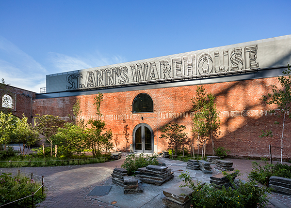 St. Ann's stage door entrance in vibrant sunlight with the St. Ann's Warehouse signage featured prominently in the garden.